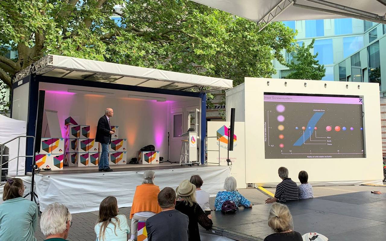 Ein Mann hält einen Vortrag auf einer Open-Air-Bühne, davor sitzen Zuschauerinnen und Zuschauer.