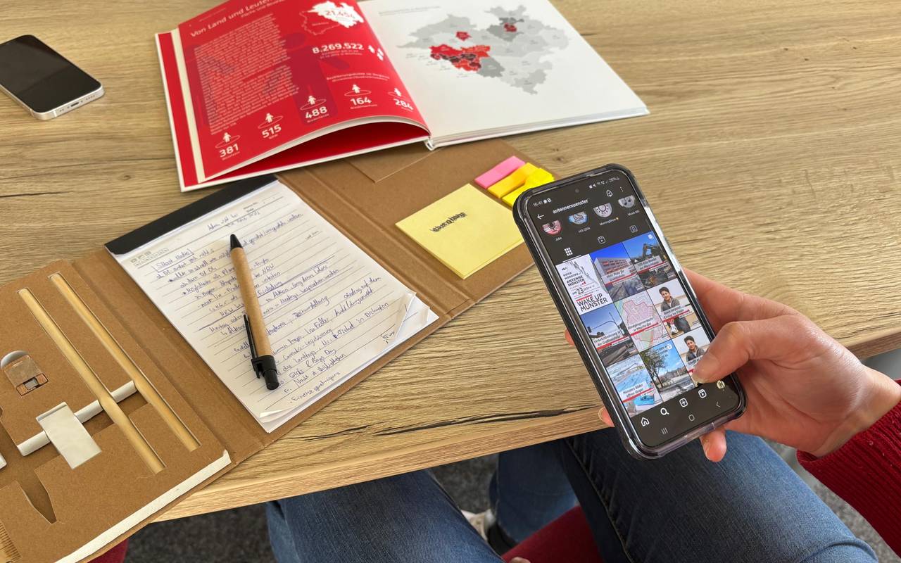 Auf dem Bild sieht man eine Person, die ein Handy in der Hand hat und an einem Tisch mit Büchern darauf sitzt.