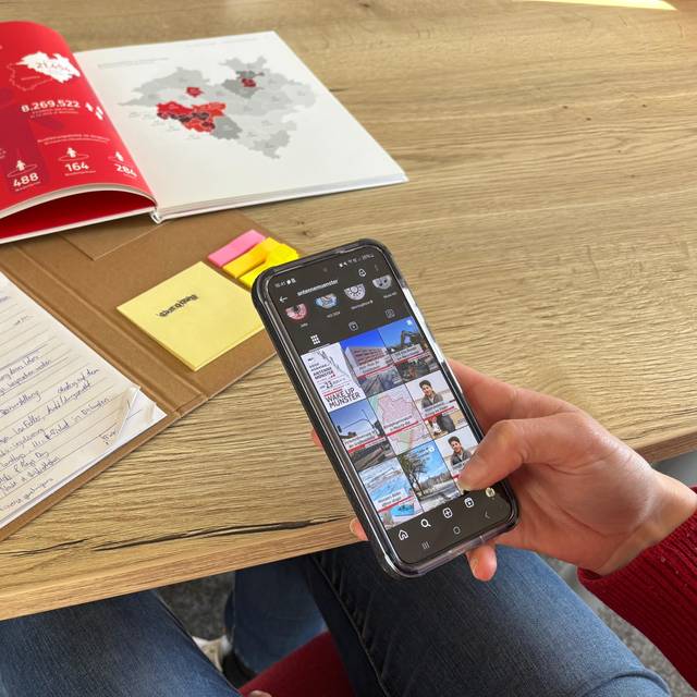 Auf dem Bild sieht man eine Person, die ein Handy in der Hand hat und an einem Tisch mit Büchern darauf sitzt.