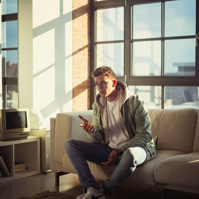 Ein junger Mann mit braunen Haaren sitzt auf einem Sofa und singt.