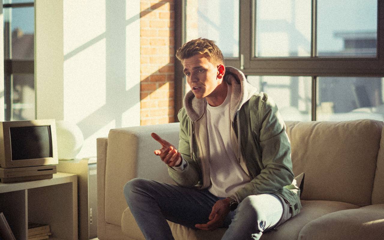 Ein junger Mann mit braunen Haaren sitzt auf einem Sofa und singt.