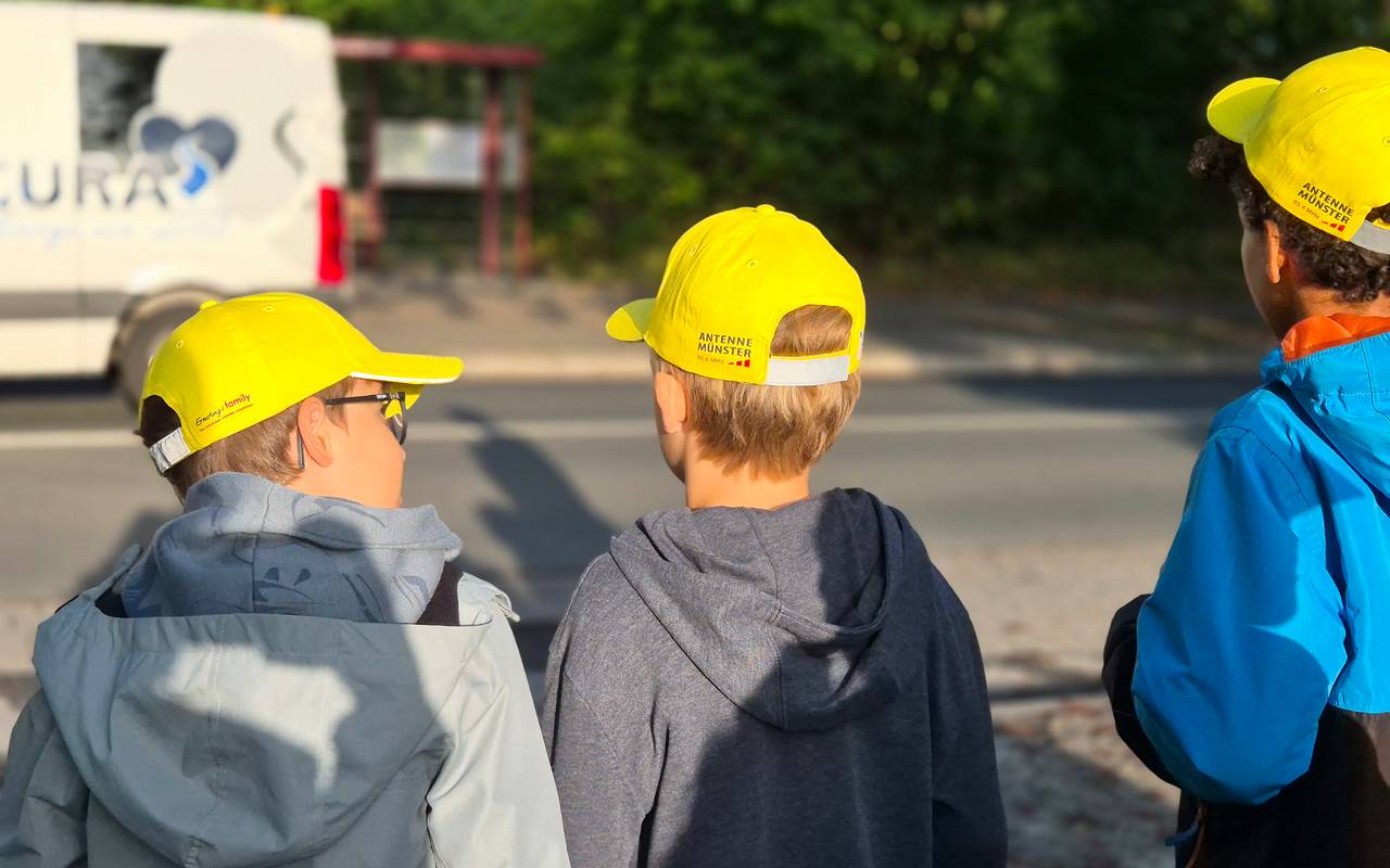 Drei Kinder mit gelben Käppis stehen an einer Straße.