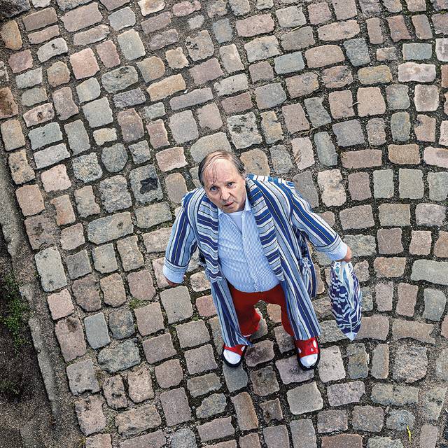 "Dittsche" steht im Bademantel, Latschen und mit Aldi-Tüte in der Hand auf einem Kopfsteinpflaster.