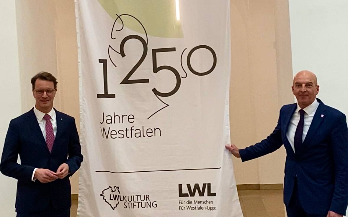 Auf der linken Seite steht Ministerpräsident Hendrik Wüst. Auf der anderen Seite steht der LWL-Direktor Dr. Georg Luhnemann. Zwischen ihnen hängt ein Banner. Darauf steht: 1250 Jahre Westfalen.