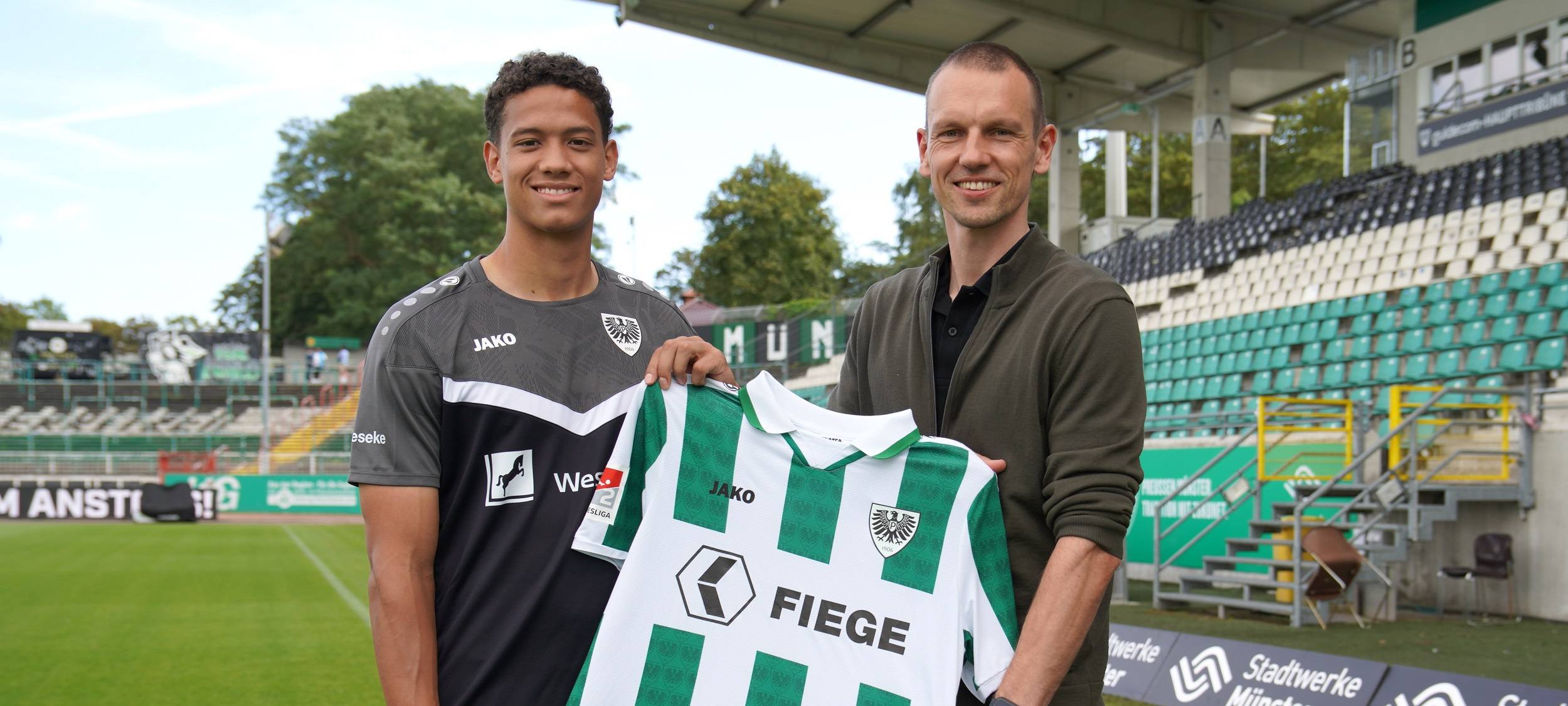 András Németh steht in einem grauen Trikot auf der linken Seite des Bildes. Ole Kittner steht neben ihm. In ihrer Mitte halten sie das weiß-grün gestreifte Trikot des SC Preußen Münster.