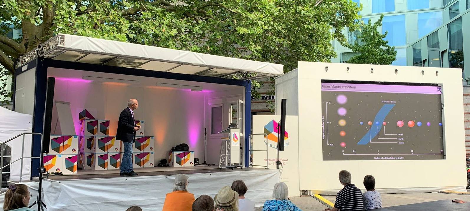 Ein Mann hält einen Vortrag auf einer Open-Air-Bühne, davor sitzen Zuschauerinnen und Zuschauer.