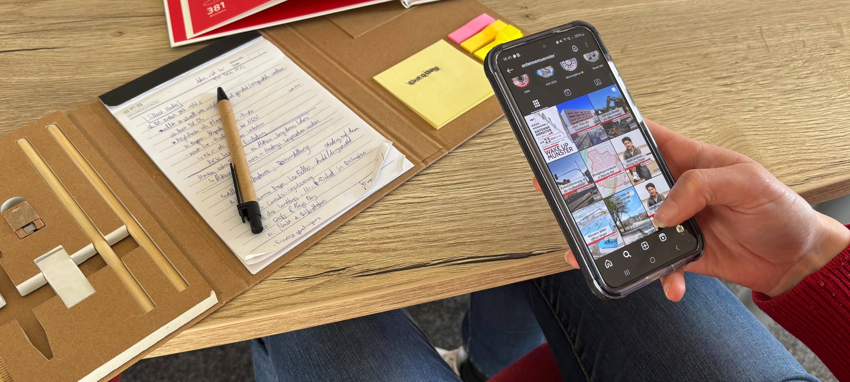 Auf dem Bild sieht man eine Person, die ein Handy in der Hand hat und an einem Tisch mit Büchern darauf sitzt.
