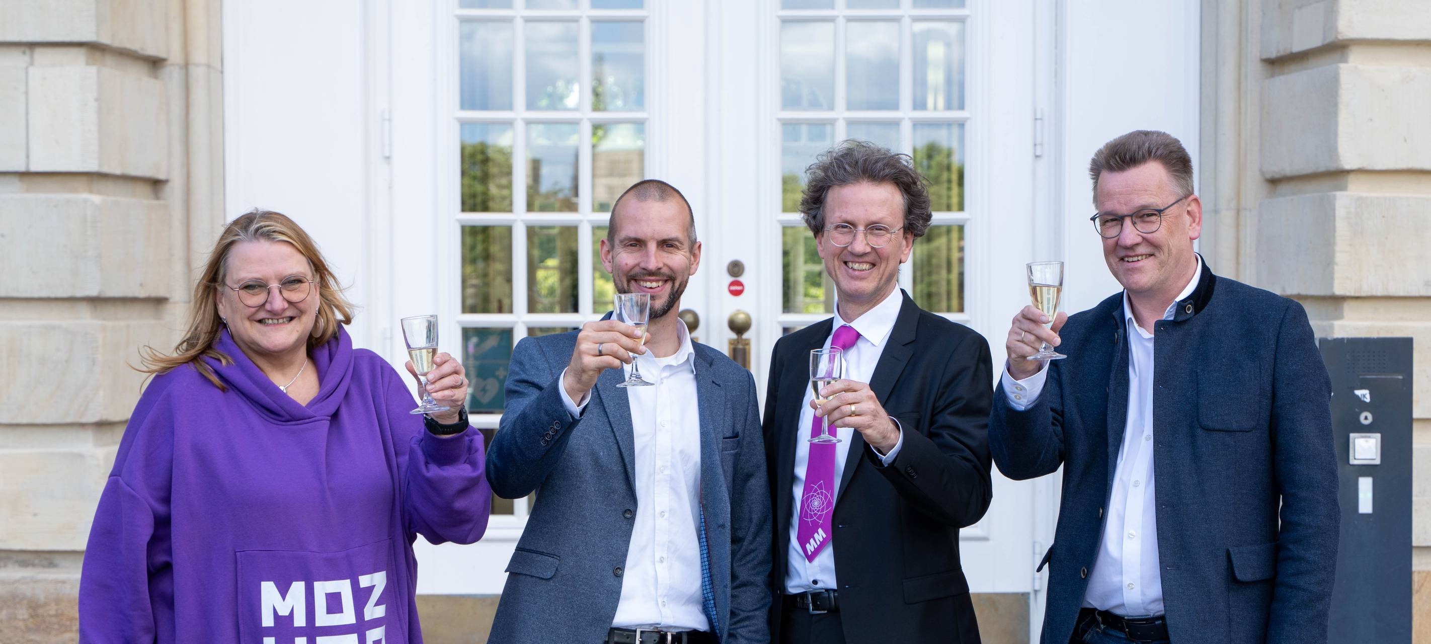 Drei Männer und eine Frau stoßen mit Sekt an.