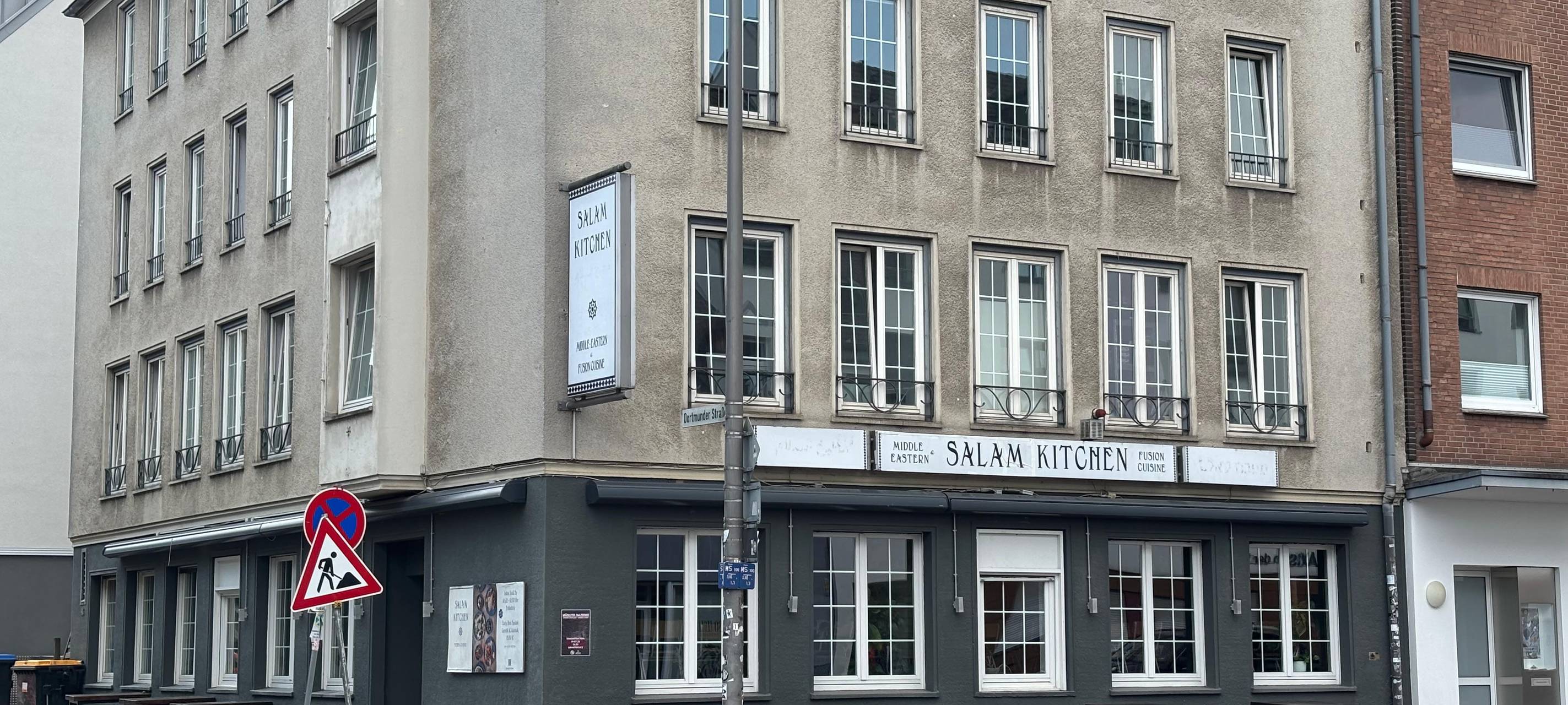 Ein Restaurant im unteren Bereich eines Wohnhauses. Es ist in schwarz gestrichen. Auf einem Schild steht der Name des Restaurants: Salam Kitchen.
