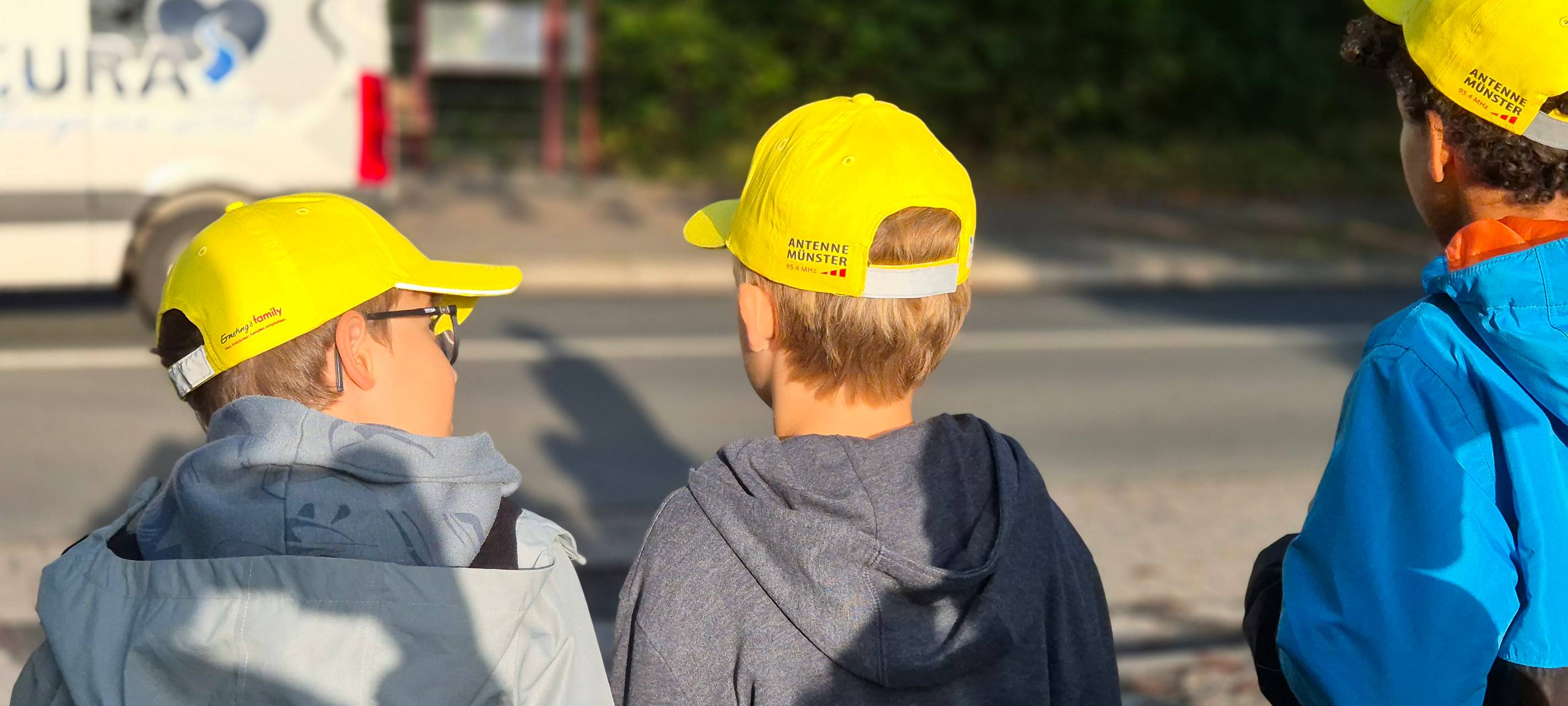 Drei Kinder mit gelben Käppis stehen an einer Straße.